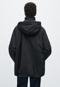 Giacca nera con cappuccio realizzata in tessuto leggero, caratterizzata da una vestibilità ampia e una superficie testurizzata, con coulisse sul cappuccio e senza chiusure visibili.