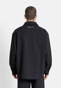 Hombre con cabello corto que lleva una camisa de manga larga negra texturizada y pantalones a juego, visto desde atrás sobre un fondo blanco.