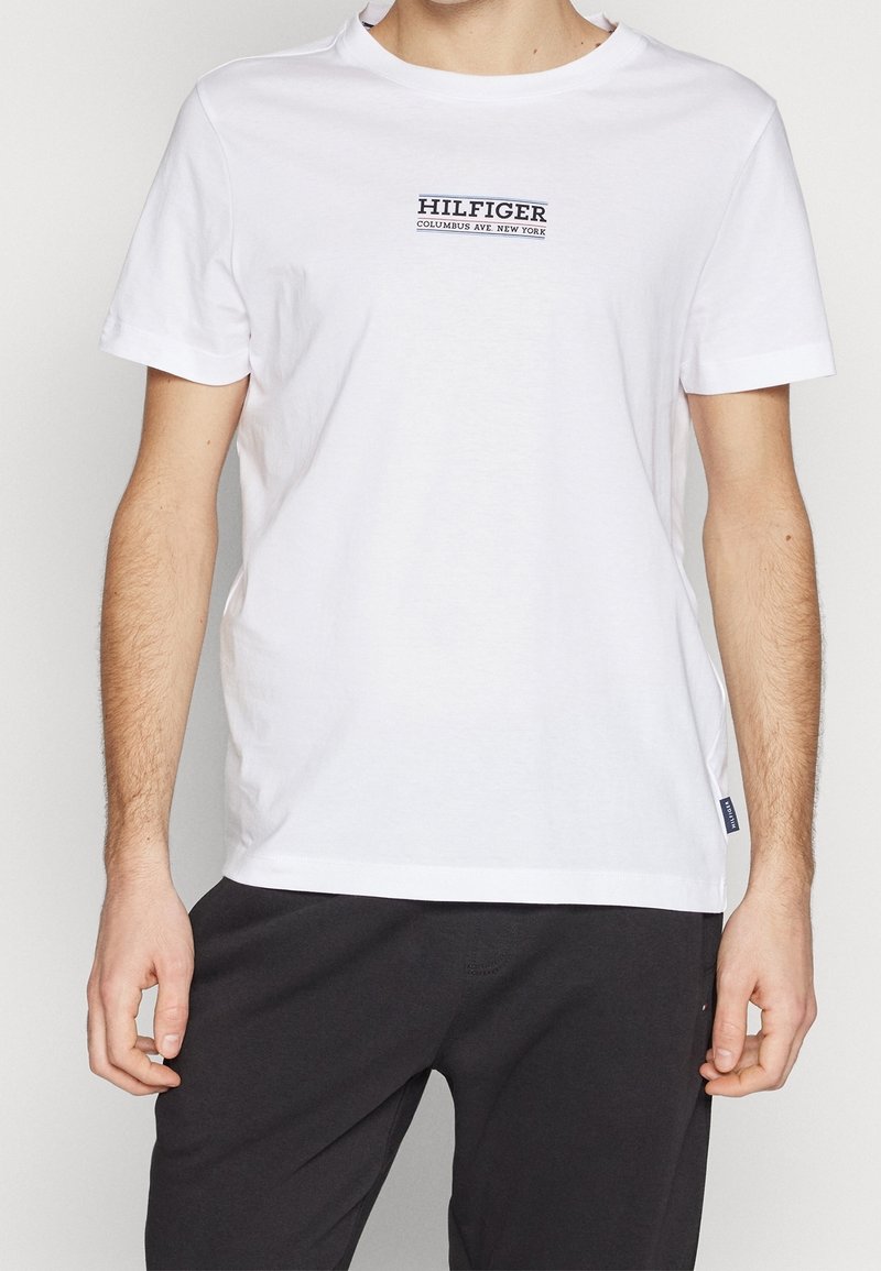 Mies, joka pukeutunut valkoiseen lyhythihaiseen T-paitaan, jossa on rintapuolella teksti "HILFIGER COLUMBUS AVE. NEW YORK", ja mustiin housuihin.