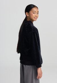 Felpa in velluto a coste nera con scollo rotondo e polsini a coste, abbinata a pantaloni larghi grigi, vista di lato.