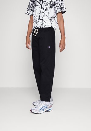 Personne portant un pantalon noir à cordon avec le logo Champion, un t-shirt graphique abstrait noir et blanc, des chaussettes blanches et des chaussures de course argentées et bleues.