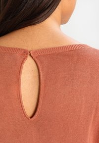 Haut en maille couleur rouille avec un col rond et une petite ouverture en forme de goutte d'eau à l'arrière. Texture lisse avec une coupe ajustée et un subtil côtes.