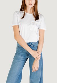 Camiseta blanca de algodón con mangas cortas, con un bolsillo adornado con cuentas ovaladas en tonos pastel. Modelo combinada con jeans azul de tiro alto.