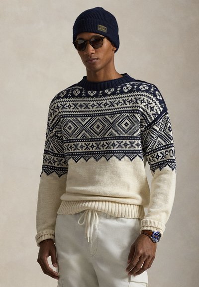Pull en maille à motifs géométriques bleu marine et crème, avec poignets et ourlet côtelés, accompagné d'un bonnet bleu marine et de lunettes de soleil.