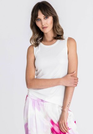 Femme aux cheveux bruns mi-longs, portant un haut blanc sans manches et un pantalon tie-dye blanc et rose, debout avec un bras croisé sur l'autre.