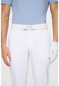 Biały pasek ze srebrną geometryczną klamrą, połączony z białymi spodniami i jasnoniebieską koszulą polo. Na prawej ręce widoczna jest biała rękawica golfowa.