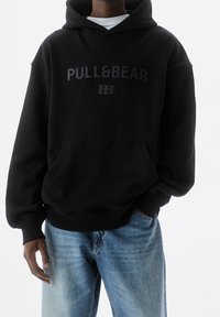 Personne portant un sweat à capuche noir Pull&Bear et un jean bleu clair, debout avec une main dans la poche, sur un fond blanc uni.