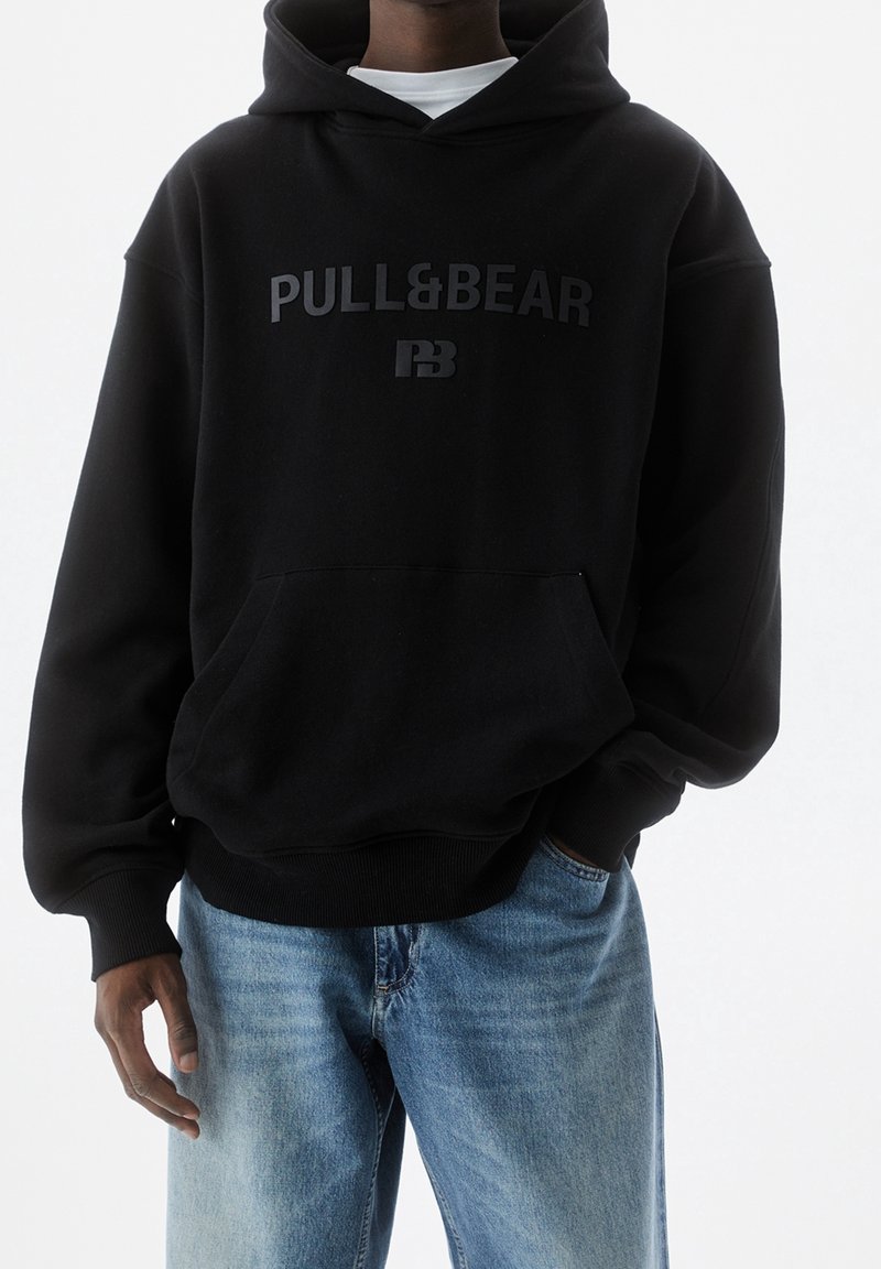 Personne portant un sweat à capuche noir Pull&Bear et un jean bleu clair, debout avec une main dans la poche, sur un fond blanc uni.