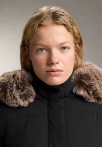 Jachetă neagră căptușită, cu guler înalt și margine mare din blană sintetică texturată, de culoare maro. Material neted, mat, cu detalii cusute.