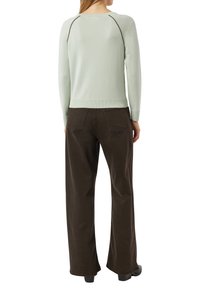 Kobieta w jasnozielonym topie z długim rękawem i ciemnobrązowych spodniach o szerokich nogawkach, stojąca tyłem z rękami swobodnie opuszczonymi wzdłuż ciała.