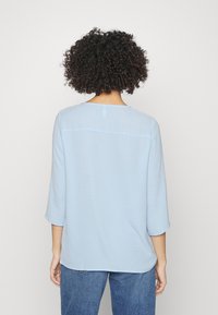 Blusa azzurro chiaro, con texture, maniche a tre quarti, scollatura rotonda e un dettaglio di cucitura orizzontale sul retro.