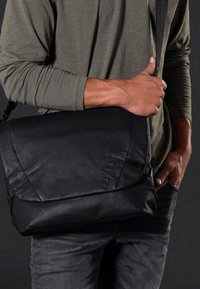 Zwarte boodschappentas gemaakt van glad materiaal, met een vlak voorflap, gestikte details en een lange verstelbare riem.