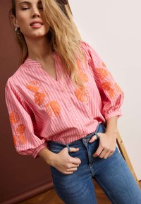 Mujer con blusa rosa abotonada con bordado de hojas naranjas y rayas verticales, combinada con vaqueros azules, de pie con las manos cerca de la cintura.