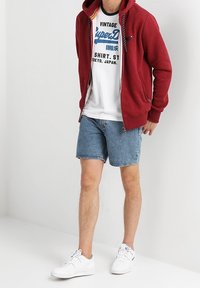 Sudadera roja con cremallera y tejido texturizado, camiseta gráfica blanca, pantalones cortos de mezclilla azul claro y zapatillas blancas con acentos sutiles.