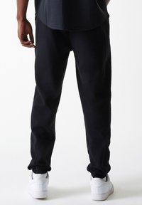 Schwarze Jogginghosen mit elastischen Bündchen, aus weichem Stoff. Getragen mit weißen Sneakers. Das Design bietet eine lockere Passform und minimale Details.