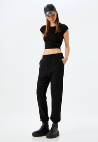 Schwarzes Crop-Top und schwarze Jogginghosen mit Kordelzug aus weicher Stoffqualität, kombiniert mit schwarzen flachen Stiefeln und einer lederähnlichen Mütze.