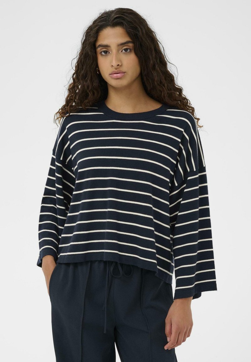 Marineblå stribet pullover med cremefarvede striber, afslappet pasform, brede ærmer, ribbet hals, tekstureret strikket stof, båret over mørke bukser.