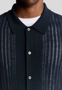 Mörkstickad polo med en strukturerad mönstring, som har vertikala ränder och fyra knäppningar i kragen i en kontrasterande färg.