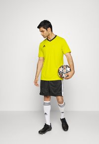 Żółta koszulka sportowa z czarnym dekoltem w kształcie V, w połączeniu z czarnymi szortami i białymi skarpetami piłkarskimi. Zawodnik trzyma wzorzystą piłkę nożną.