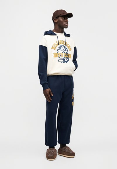 Σκούρο μπλε αθλητικό σετ με κρεμ φούτερ με κουκούλα που φέρει το κείμενο «Billionaire Boys Club». Καφέ παντόφλες με λεπτομέρειες κεντήματος με λουλούδια.