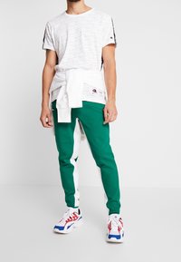 Man som bär en vit t-shirt med logotyp, en vit sweatshirt band över midjan, gröna och vita träningsbyxor, samt vita sneakers med röda och blå accenter.