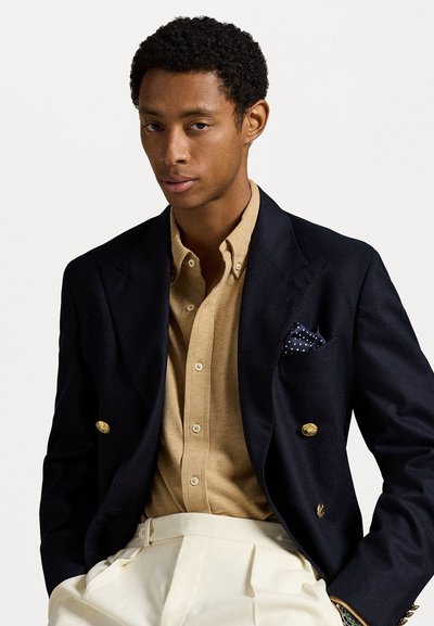 Blazer azul-marinho de dois botões com botões dourados sobre uma camisa castanho-claro. Calças brancas plissadas e um lenço de bolso azul-marinho com pontos brancos.