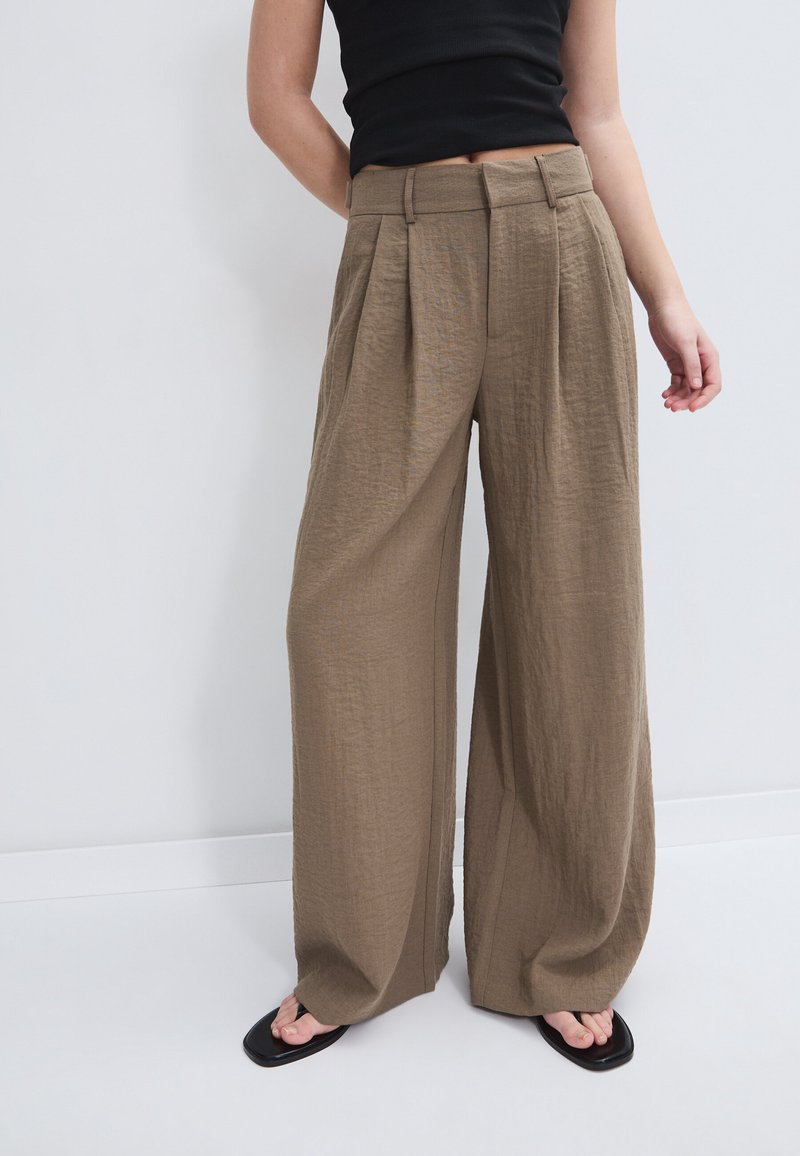 Femme portant un pantalon marron taille haute à jambes larges avec des plis, associé à un haut noir sans manches et des sandales noires à bout carré.