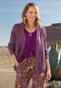 Cardigan in maglia leggera viola sopra una blusa viola con scollatura a chiave, abbinato a pantaloni floreali colorati e a fantasia. Accessori dorati.