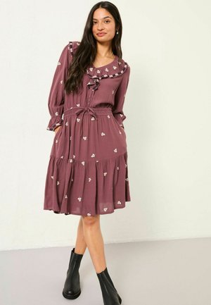 Mauve Kleid mit langen Ärmeln, floraler Stickerei, Rüschensaum und Kordelzugtaille. Ausgestellter Saum und kombiniert mit schwarzen Knöchelstiefeln.