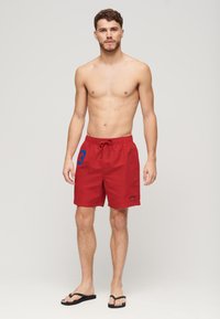 Shorts de bain rouges avec une taille à cordon, présentant un numéro "3" bleu sur le côté et un petit logo près de l'ourlet.