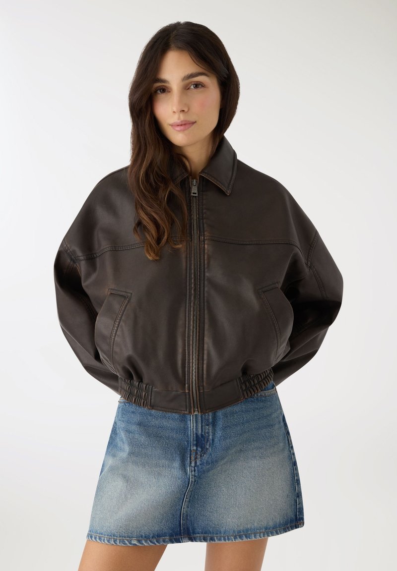 Donna con lunghi capelli castani che indossa una giacca con zip in pelle marrone scuro e una gonna di jeans blu sbiadito su uno sfondo uniforme.