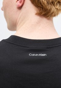 Άτομο με ανοιχτόχρωμο δέρμα και κόκκινα μαλλιά που φοράει μαύρο πουκάμισο με μικρό λευκό λογότυπο "Calvin Klein" στο πίσω μέρος του λαιμού.