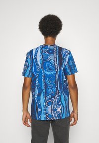 Person med kort lockigt hår bär en blå mönstrad kortärmad skjorta och mörkgrå byxor, stående mot en vit bakgrund.