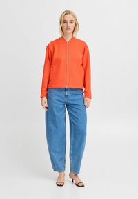 Oranžová mikina na zips so bočnými vreckami, kombinovaná s voľnými modrými džínami. Textúrovaná tkanina, neformálny dizajn a dlhé rukávy. Čierne sandále na podpätku.
