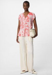 Top a maniche corte con stampa floreale in corallo e bianco, abbinato a pantaloni larghi crema. Il modello tiene una piccola borsa beige. Scarpe punta in crema.
