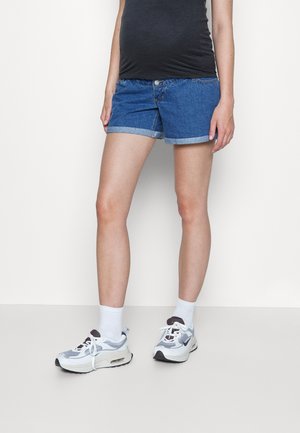 Persoon draagt blauwe opgerolde denimshorts, witte sokken en witte sportschoenen, staand tegen een effen lichte achtergrond.