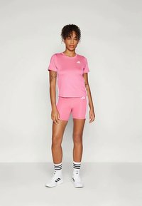 Roosa Adidas spordikomplekt, mis sisaldab lühikeste varrukatega särki ja sobivaid lühikesi pükse, valgete triipude ja logo aktsentidega. Mudel kannab valgeid tossusid ja triibulisi sokke.