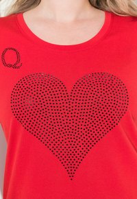 Rød bomulds T-shirt med et stort design af sorte rhinsten i form af et hjerte samt et lille "Q" i sorte rhinsten øverst til venstre.