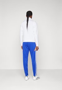 White zip-up jacket with stand collar; blue sweatpants featuring orange accents and white side stripes; model facing away, wearing sneakers.