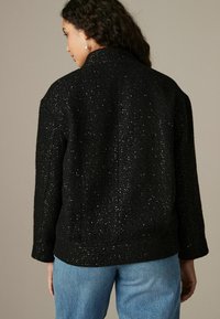 Svart texturerad jacka med lös passform, med metalliska detaljer, hög krage och långa ärmar. Bäraren har på sig ljusblå jeans.