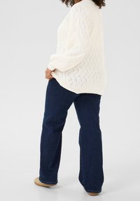 Crème gebreide trui met een textuurpatroon, gecombineerd met donkerblauwe flare jeans en lichtbruine schoenen. Focus op comfort en casual ontwerp.
