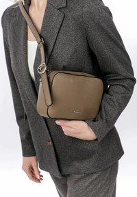 Tan leather crossbody bag with a textured surface, black zipper, and silver hardware. Compliments a gray suit jacket.