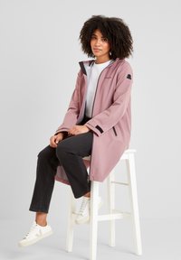 Light pink waterproof jacket with a hood, black accents, and logo. Worn over a white top; paired with dark jeans and white sneakers.