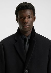 Black wool coat with a notched collar, single button closure, and smooth texture. Paired with a black tie and shirt.