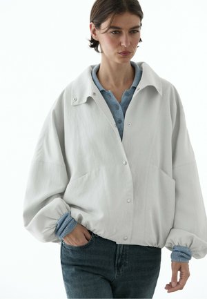 Donna che indossa una giacca larga grigio chiaro con bottoni sopra un maglione blu lavorato a maglia, con le mani nelle tasche di jeans scuri, guardando leggermente verso il basso e di lato.