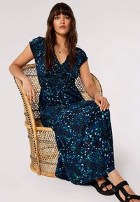 Maxikleid mit floralem Muster in Blau- und Grüntönen; kurze Ärmel, geraffter Oberteil und fließender Stoff; kombiniert mit schwarzen Riemchensandalen.