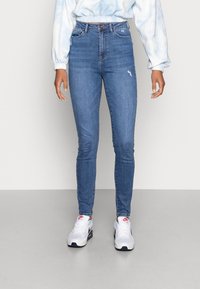 Jeans ajustados en denim azul medio con un ligero desgaste y roturas. Diseño de cintura alta con bolsillos frontales y cierre de botón.