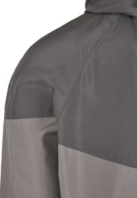 Närbild av en grå tvåfärgad jacka med en axel där sömmarna är synliga och en huva vid övre kanten.