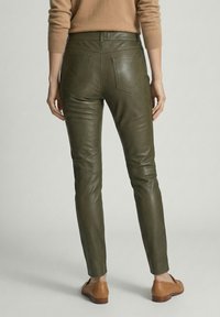 Vrouw staat met haar rug naar de camera gekeerd en draagt olijfgroene leren broek, een beige trui en lichtbruine leren instappers op een lichte achtergrond.