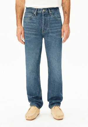 Man draagt medium blauw rechte spijkerbroek en beige mocassins, staand tegen een witte achtergrond.
